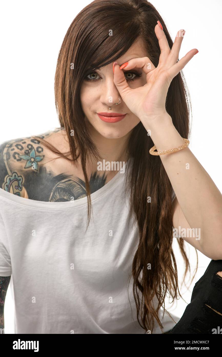 Jolie jeune femme avec de longs cheveux brunette, un T-shirt blanc et des tatouages frappants sur un fond blanc. Le pouce et l'index forment un symbole OK Banque D'Images