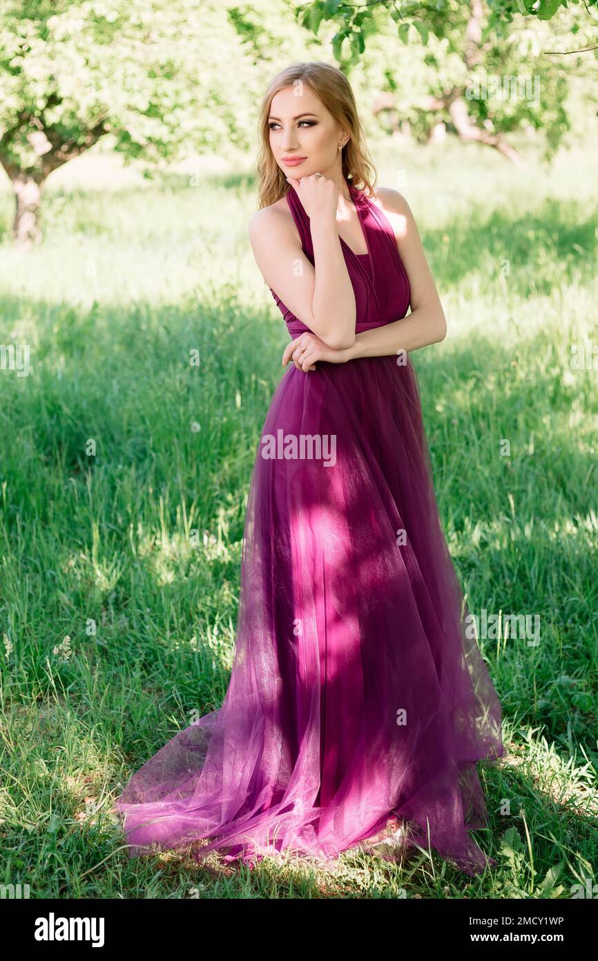 Une jeune femme d'apparence modèle se tient dans un jardin vert de printemps dans la nature dans une belle robe pourpre et pose regardant dans la distance. Banque D'Images