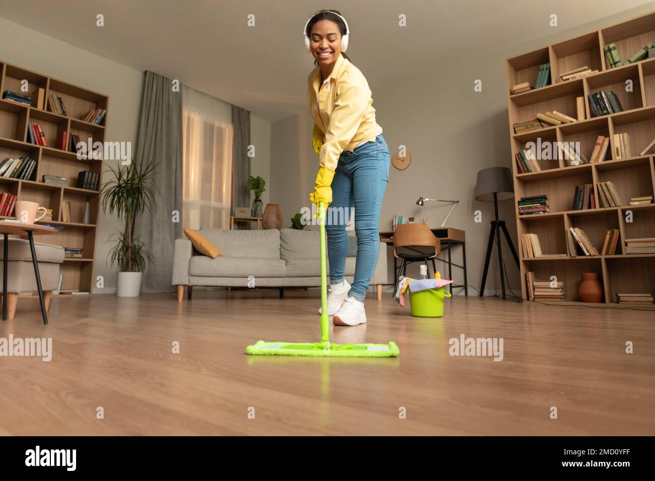 Jeune femme noire lavant le sol dans le salon en utilisant la vadrouille et en écoutant de la musique dans les écouteurs, appréciant la routine de nettoyage Banque D'Images