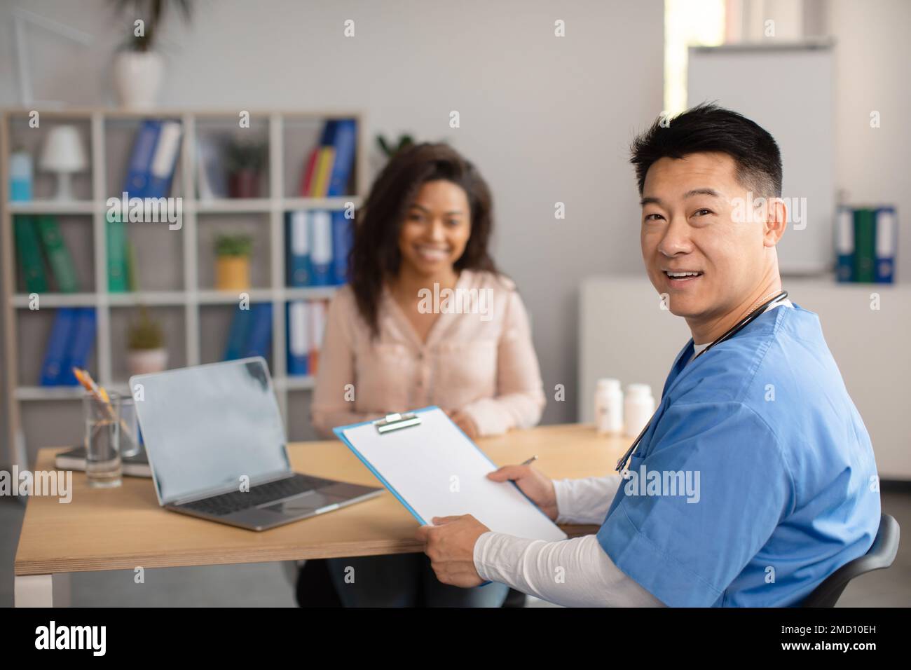 Un médecin coréen souriant d'âge moyen en uniforme consulte une jeune femme afro-américaine à l'intérieur du bureau de la clinique Banque D'Images