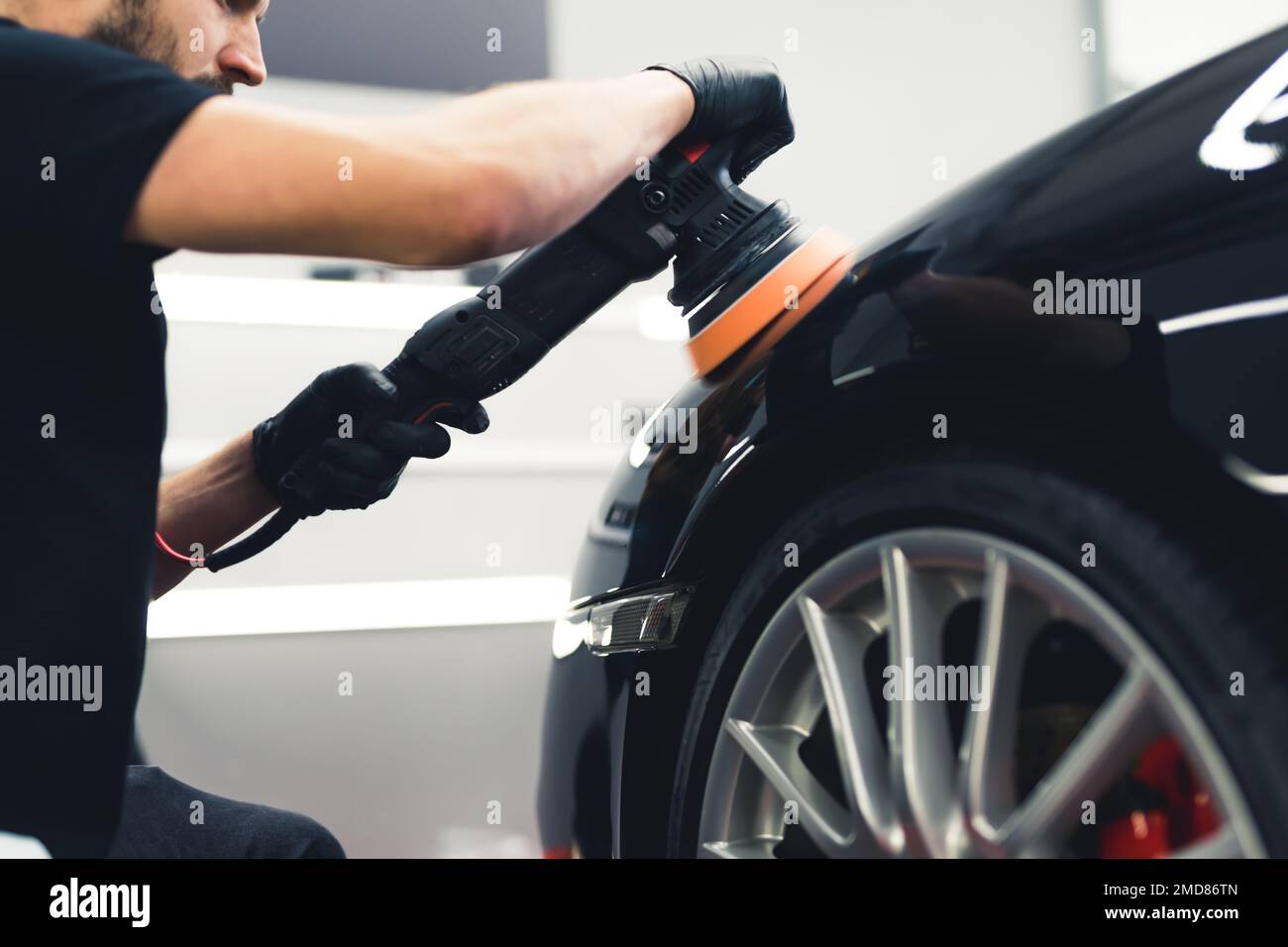 Vue à angle bas d'une voiture professionnelle détaillant un ouvrier à l'aide d'un polisseur orbital pour polir la peinture noire de la voiture. Roue et pneu dans le cliché. Photo de haute qualité Banque D'Images