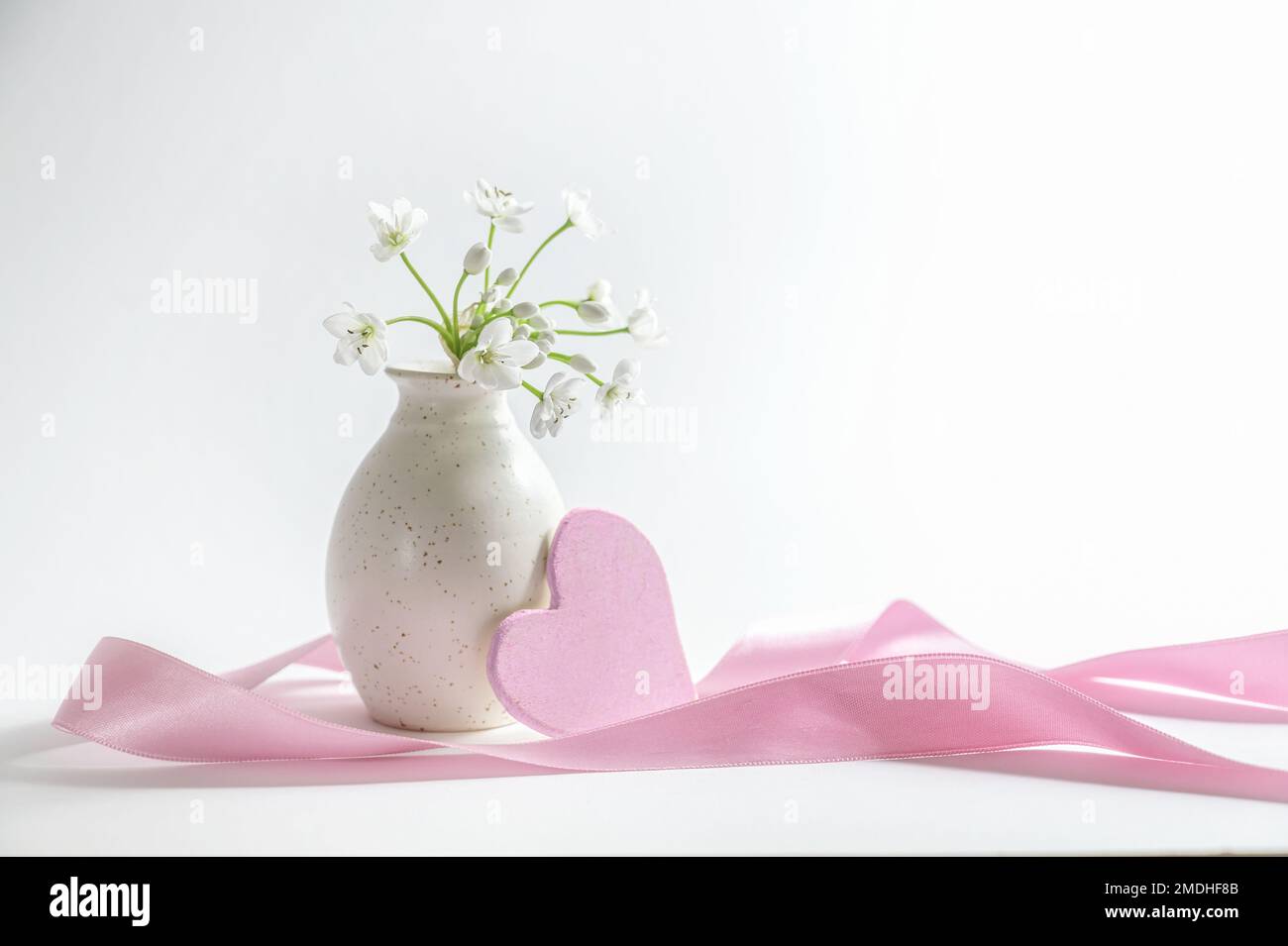 Vase avec petites fleurs blanches, coeur en bois rose et un long ruban sur fond blanc gris, symbole d'amour romantique, carte de vœux de vacances, espace copie Banque D'Images