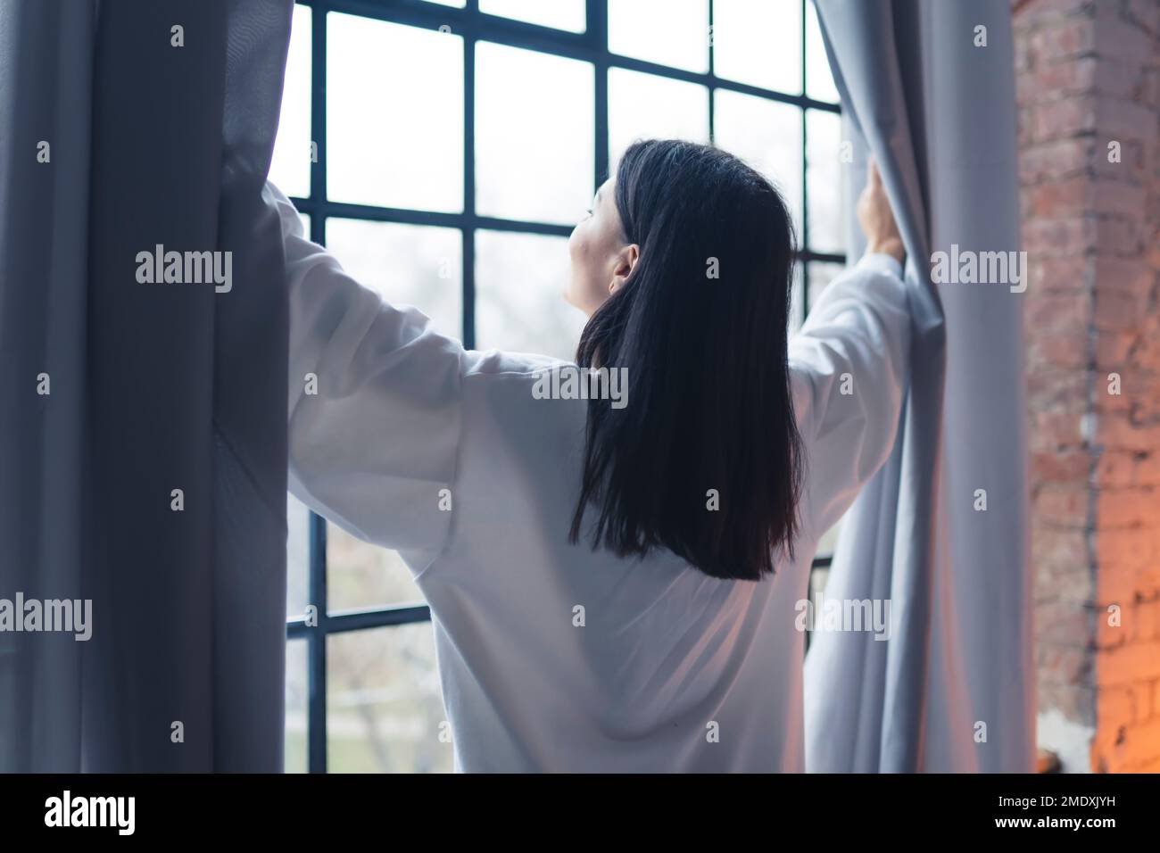 Vue arrière d'une femme ouvrant des rideaux de fenêtre et regardant à l'extérieur. Pas de face. Photo de haute qualité Banque D'Images