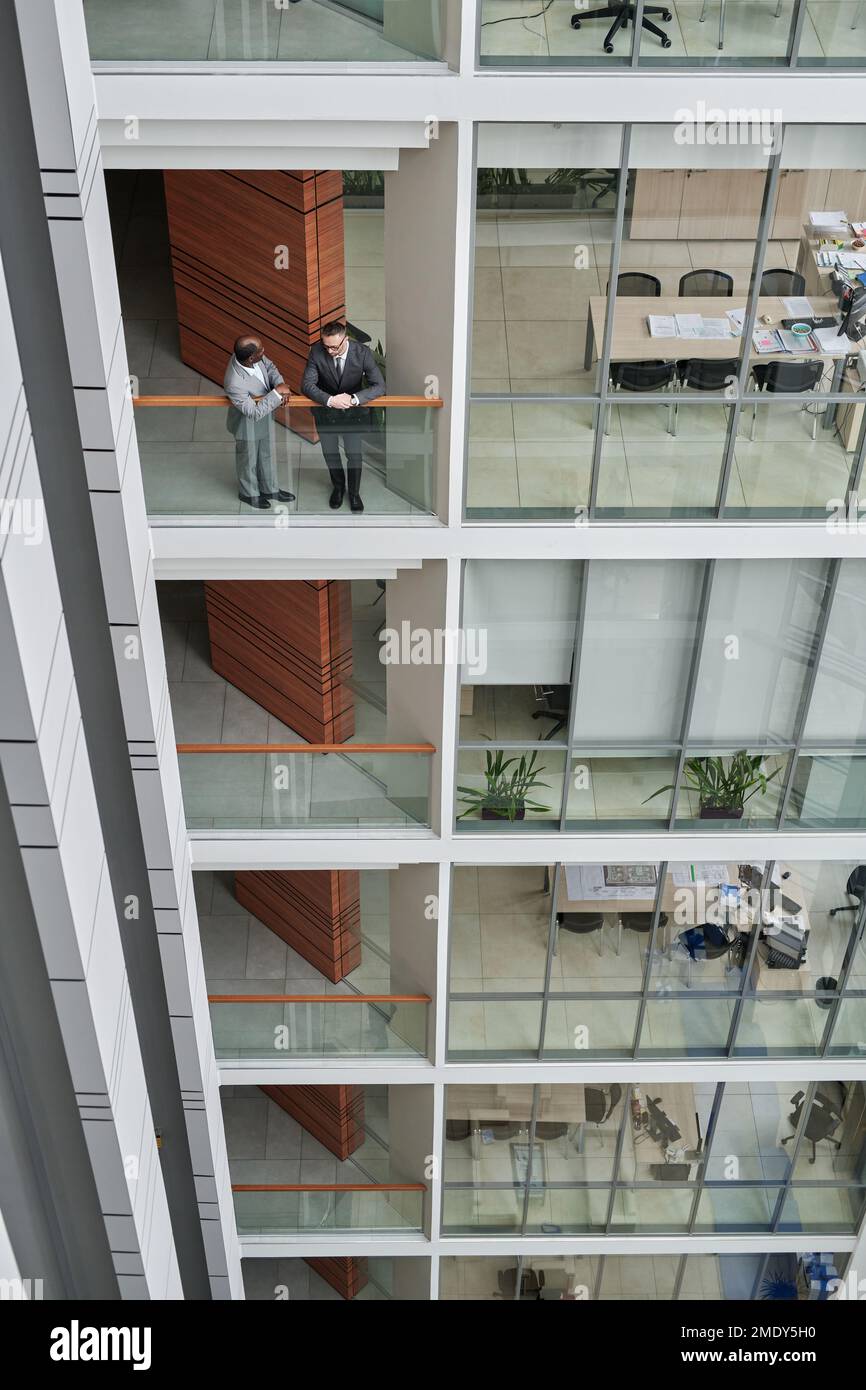 Photo ci-dessus de l'intérieur d'un bâtiment de plusieurs étages avec des bureaux à espace ouvert derrière de grandes fenêtres et deux hommes d'affaires debout près de rampes lors de réunions Banque D'Images