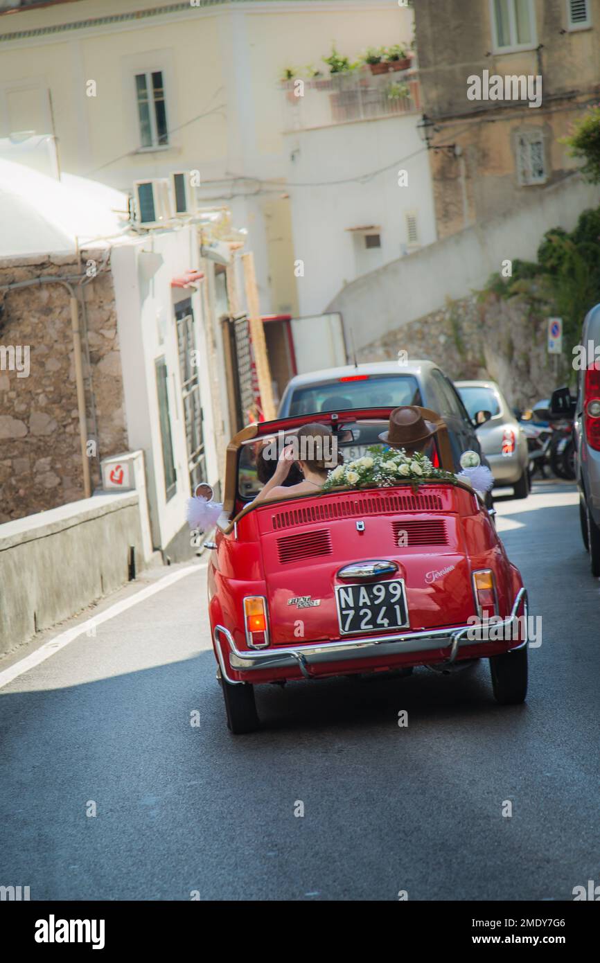 Jeune couple assis à l'arrière d'une voiture Fiat rouge décapotable vintage décorée de fleurs de mariage, Positano, Italie. Banque D'Images