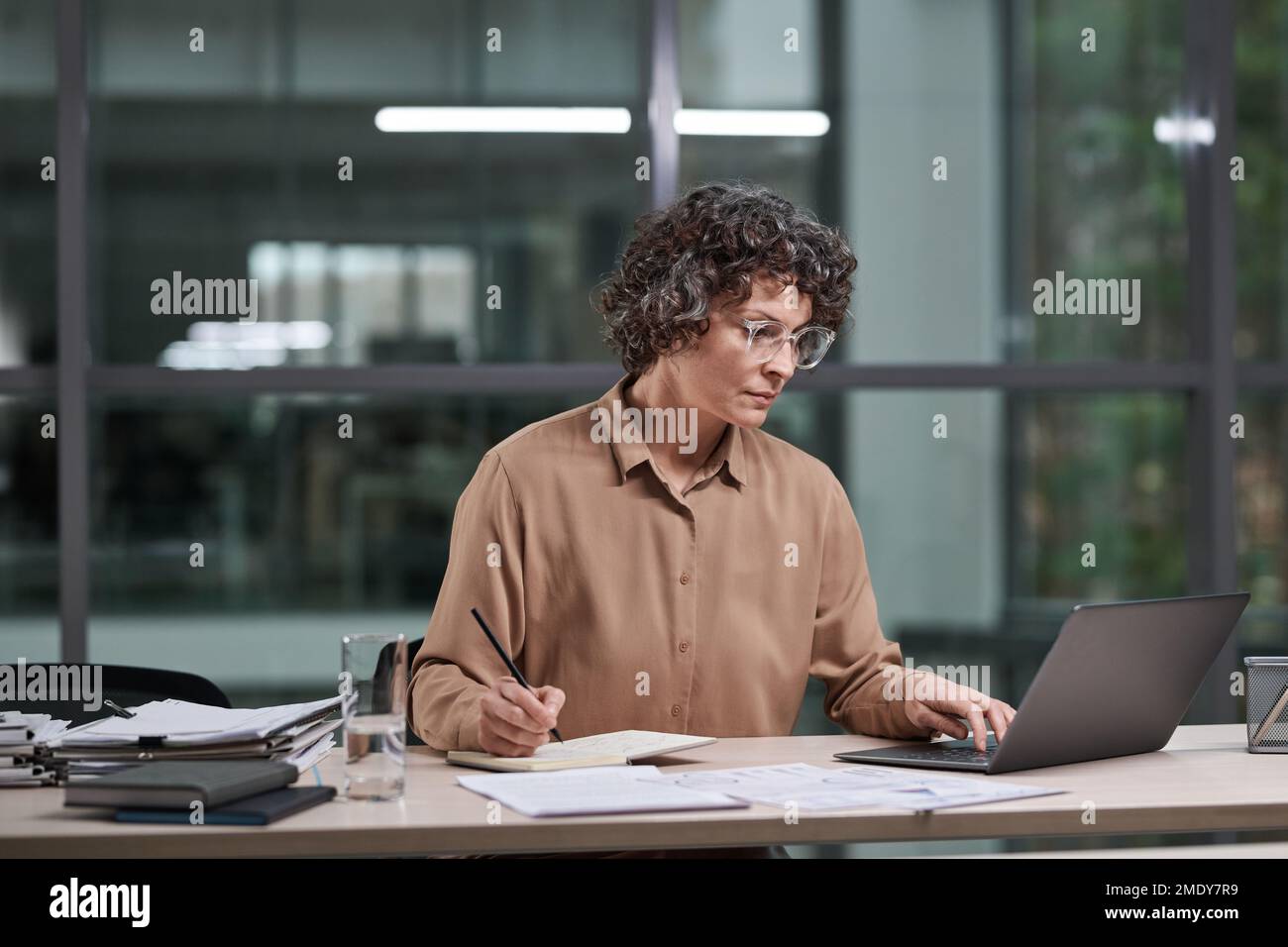 Une femme expérimentée dans un chemisier beige travaille sur un projet d'entreprise tout en étant assise sur le lieu de travail devant un ordinateur portable et en analysant les données Banque D'Images