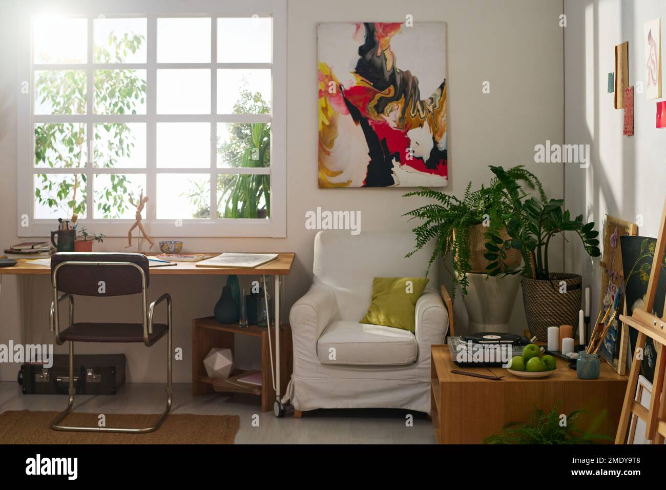 Lieu de travail de l'artiste moderne par fenêtre de studio spacieux avec table, chaise, fauteuil, peinture abstraite sur le mur et plantes vertes Banque D'Images