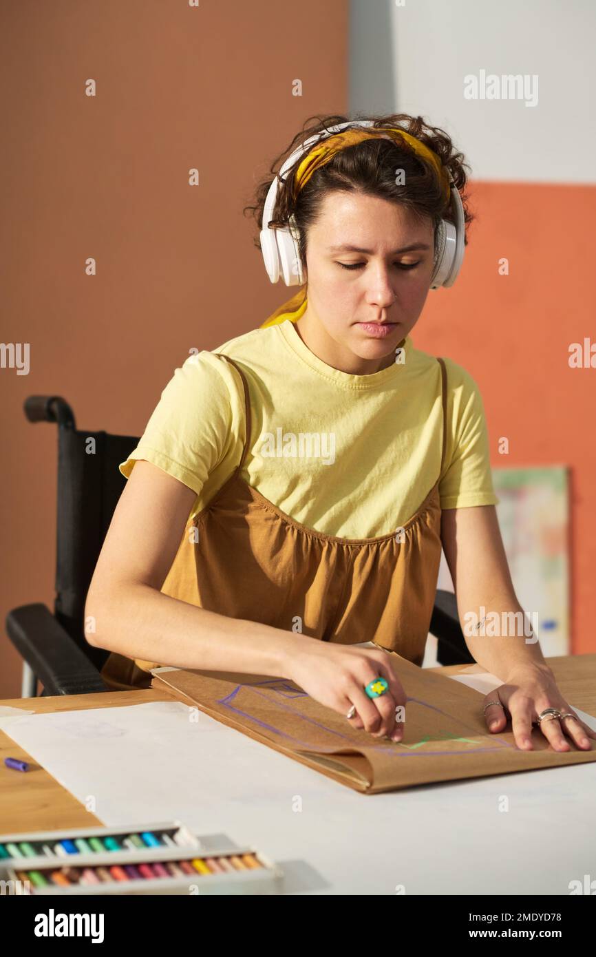 Jeune artisan sérieux avec un handicap écoutant de la musique dans un casque et esquissant avec des crayons sur une feuille de papier Banque D'Images