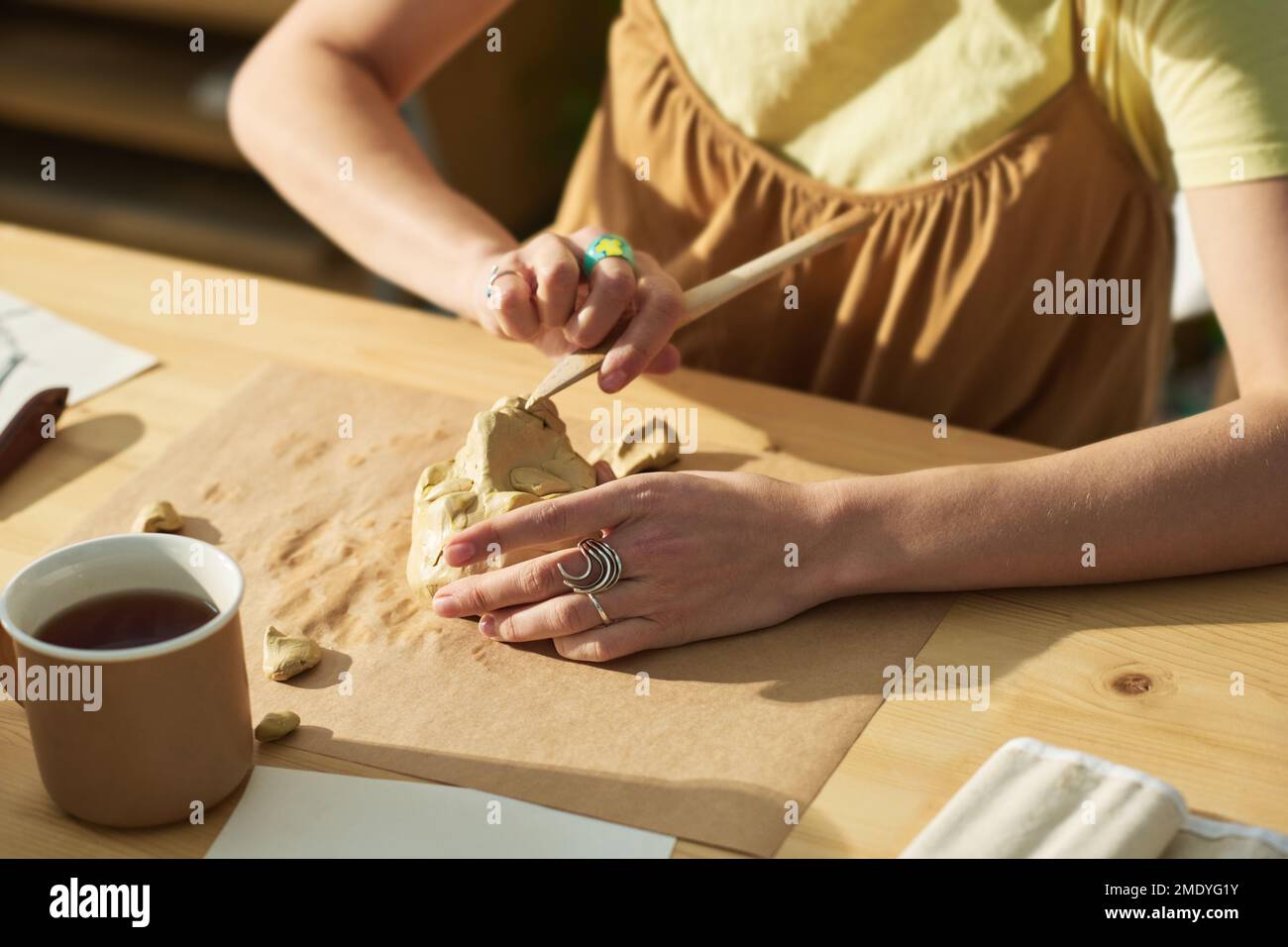 Mains de jeunes artisans créatifs avec outil en bois créant une sculpture en argile sur feuille de papier tout en étant assis sur le lieu de travail en studio Banque D'Images