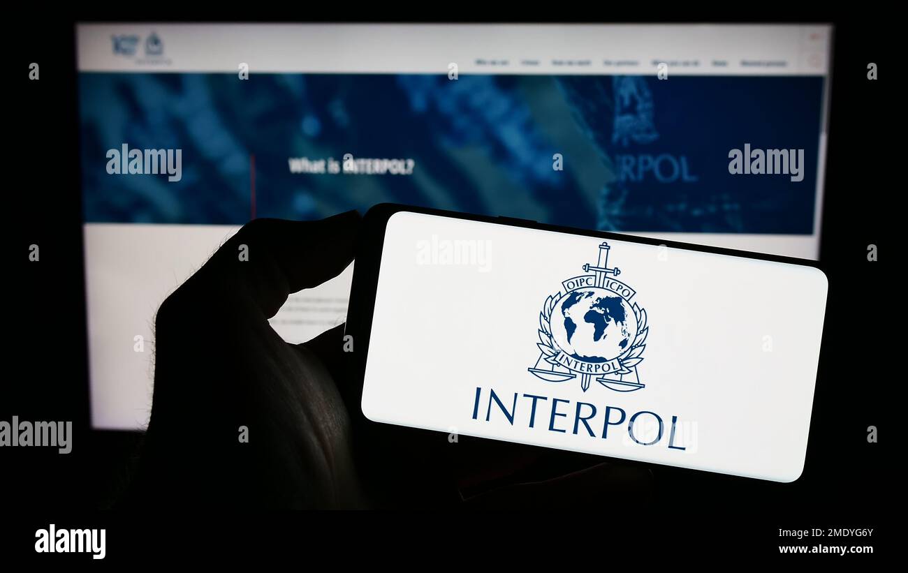 Personne tenant un smartphone avec le logo de l'Organisation internationale de police criminelle (OIPC) à l'écran devant le site Web. Mise au point sur l'affichage du téléphone. Banque D'Images