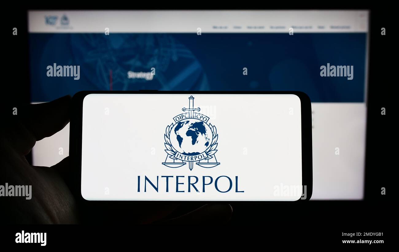 Personne tenant un téléphone portable avec le logo de l'Organisation internationale de police criminelle (OIPC) à l'écran en face de la page Web. Mise au point sur l'affichage du téléphone. Banque D'Images