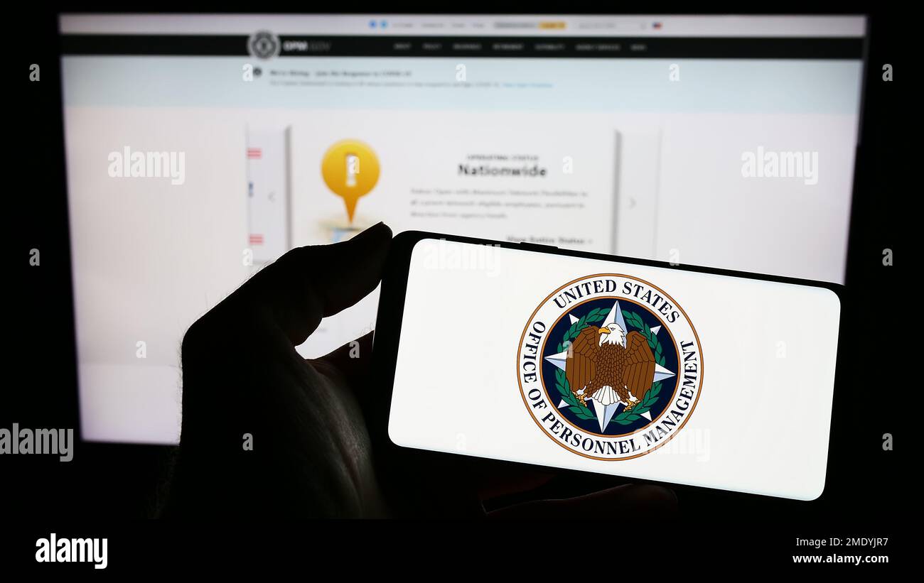 Personne tenant un téléphone portable avec le sceau de l'Office of personnel Management (OPM) des États-Unis sur l'écran en face de la page Web. Mise au point sur l'affichage du téléphone. Banque D'Images