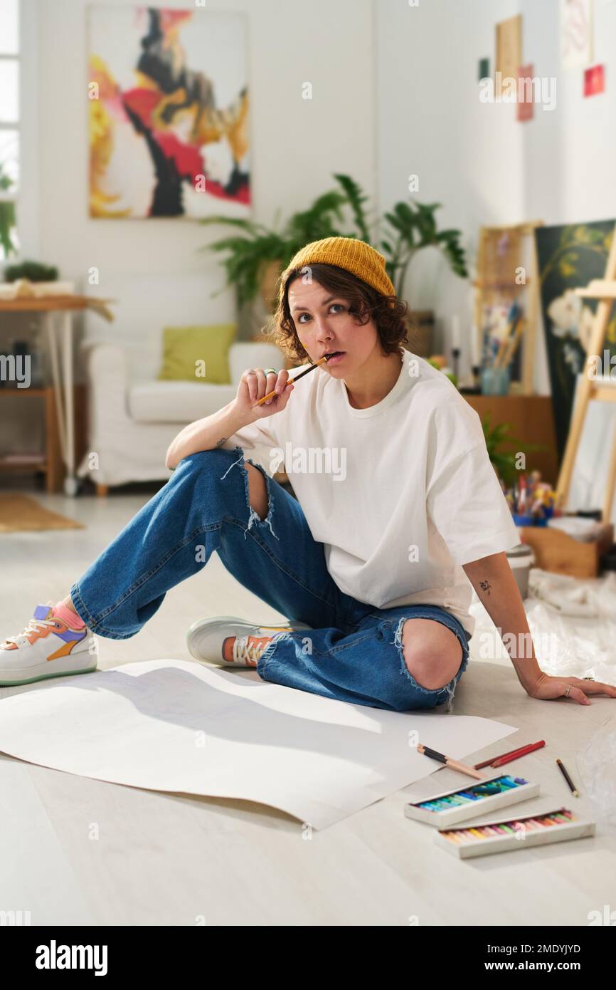 Jeune artisan pensif ou artiste en vêtements décontractés assis sur le sol entre les papiers et les fournitures pour dessiner et regarder l'appareil photo Banque D'Images