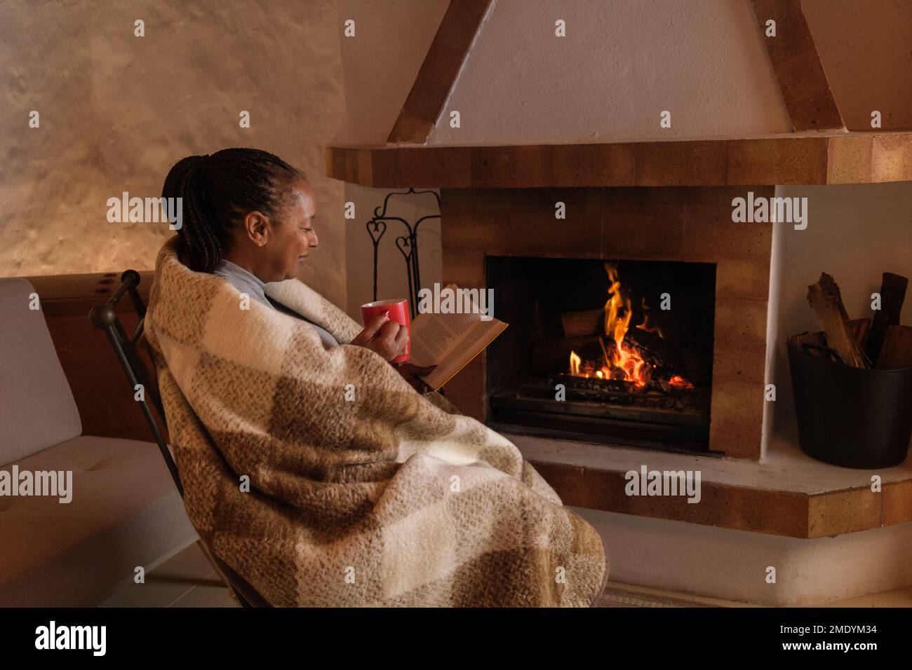 Une femme âgée lisant détendue avec une tasse de café à côté de la cheminée en hiver. Concept : retraite, repos, détente et hiver Banque D'Images