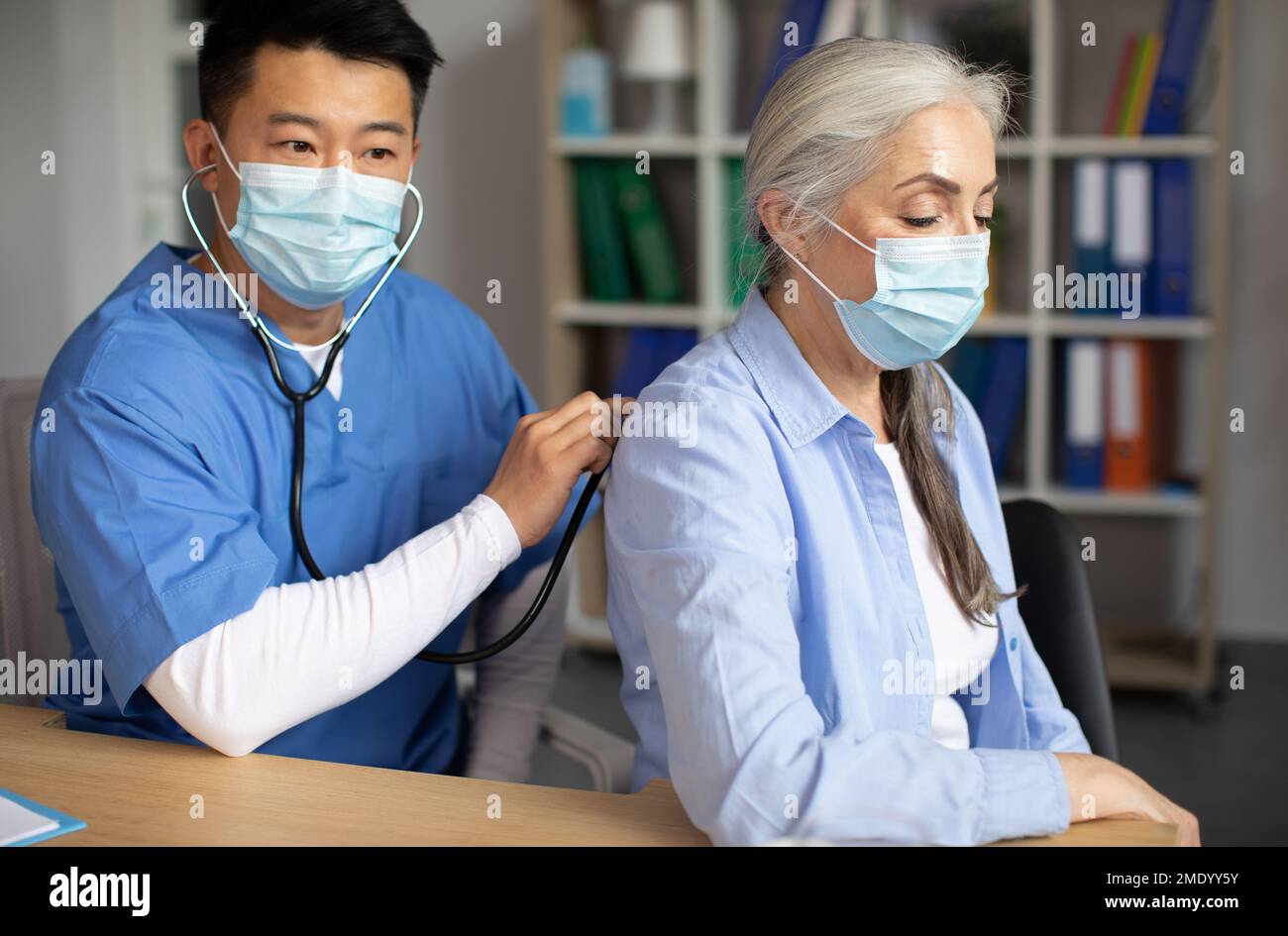 Grave homme coréen adulte médecin en masque de protection écoute avec stéthoscope à la respiration de la vieille dame européenne Banque D'Images