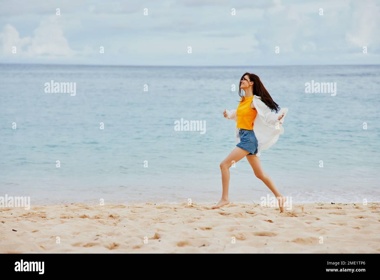 Une femme court le long de la plage en vêtements d'été sur le sable dans un t-shirt jaune et un short en Jean blanc shirt vol cheveux vue sur l'océan Banque D'Images