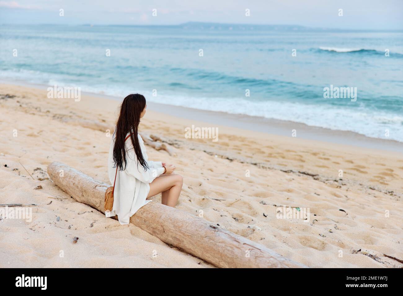 Une femme avec de longs cheveux mouillés après la natation s'assoit sur le sable avec son dos à la caméra et regarde le coucher de soleil par l'océan sur l'île de Bali Banque D'Images