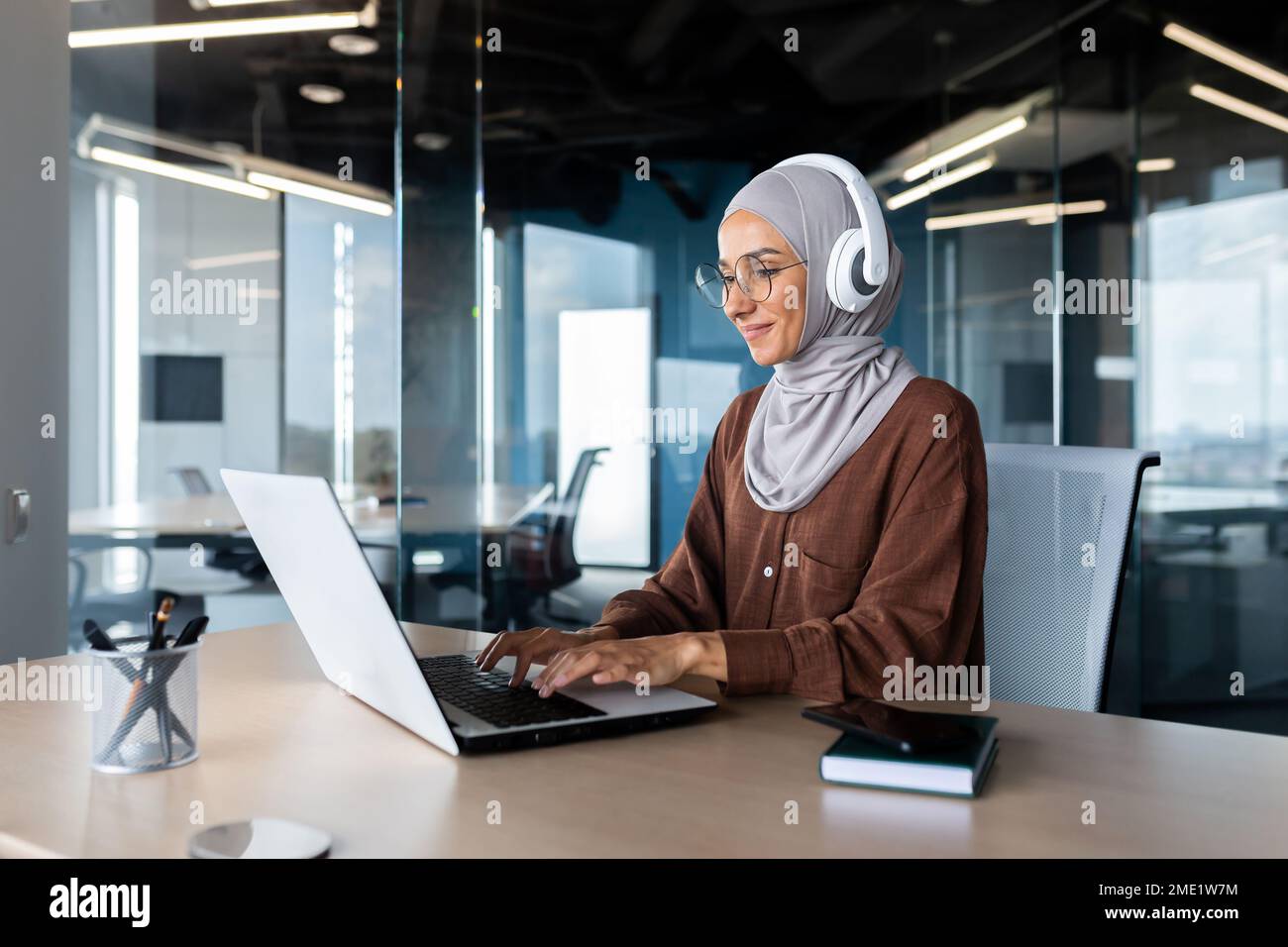 Formation en ligne. Jeune étudiante en hijab et casque, assise au bureau à la table, étudiant à distance avec un ordinateur portable. Saisie sur le clavier.sourire. Banque D'Images