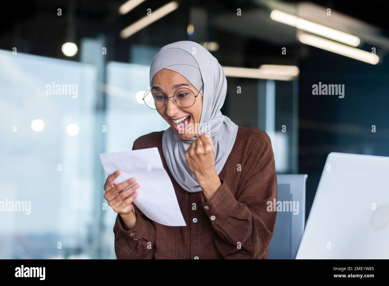 Jeune femme heureuse étudiante dans hijab. Assis à la table, tient une lettre, a reçu un avis d'admission à l'université, les résultats de l'examen. Célèbre, crie, montre le succès avec sa main. Banque D'Images