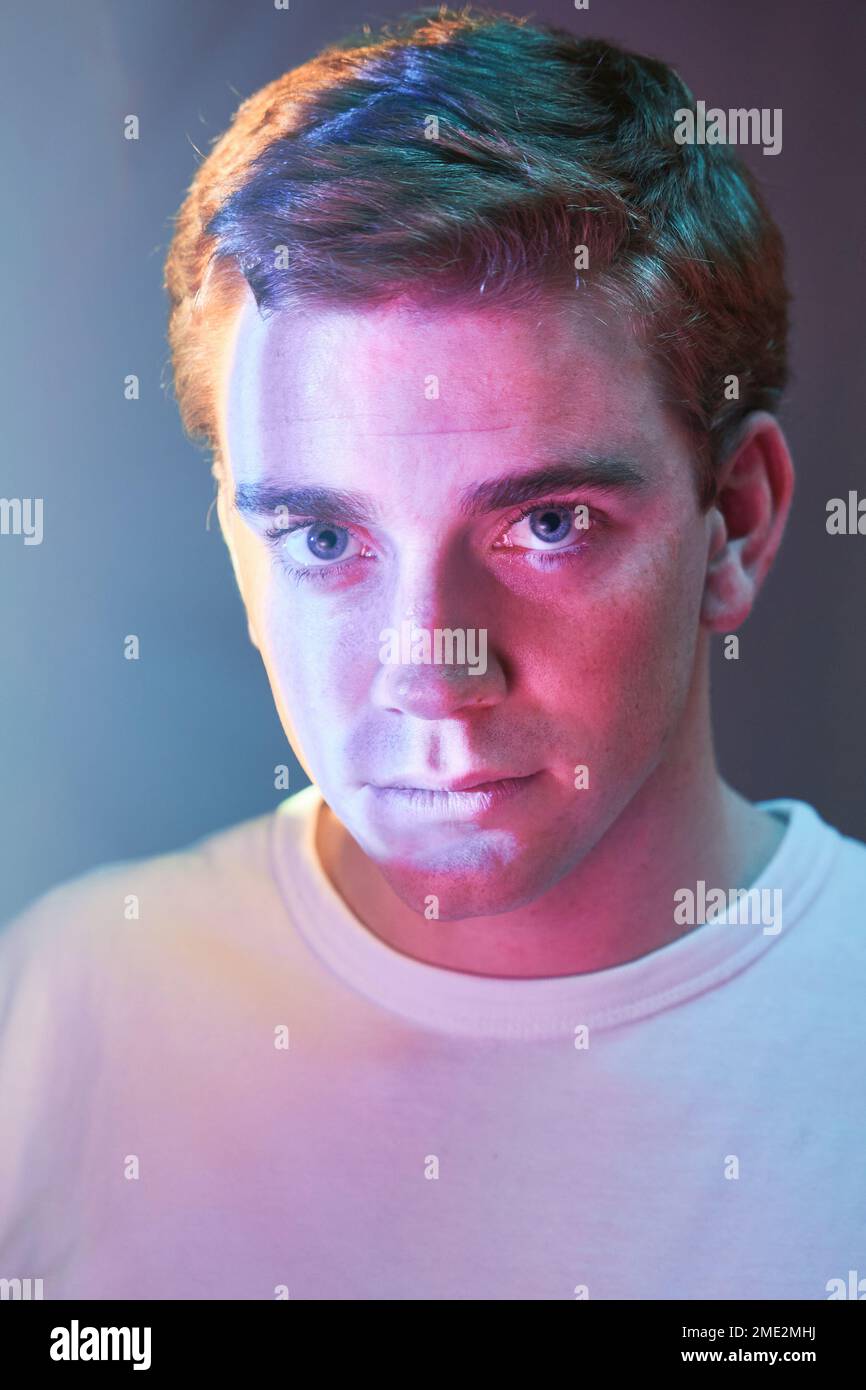 Portrait d'un jeune homme calme avec des cheveux courts portant un t-shirt blanc regardant l'appareil-photo tout en se tenant dans la lumière de néon en studio Banque D'Images