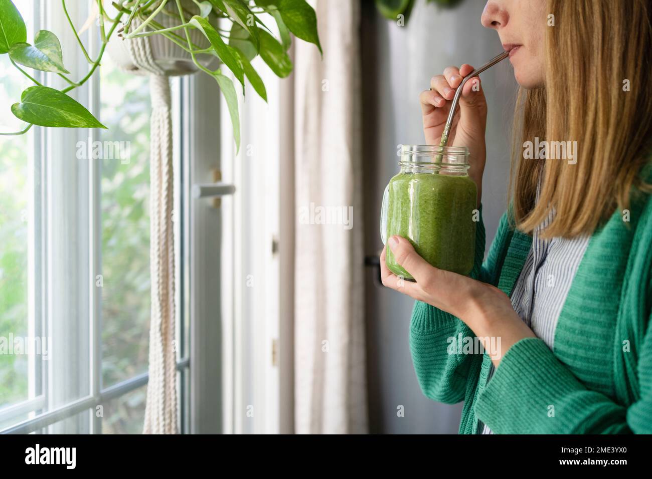 Femme buvant du smoothie vert debout près de la fenêtre à la maison Banque D'Images