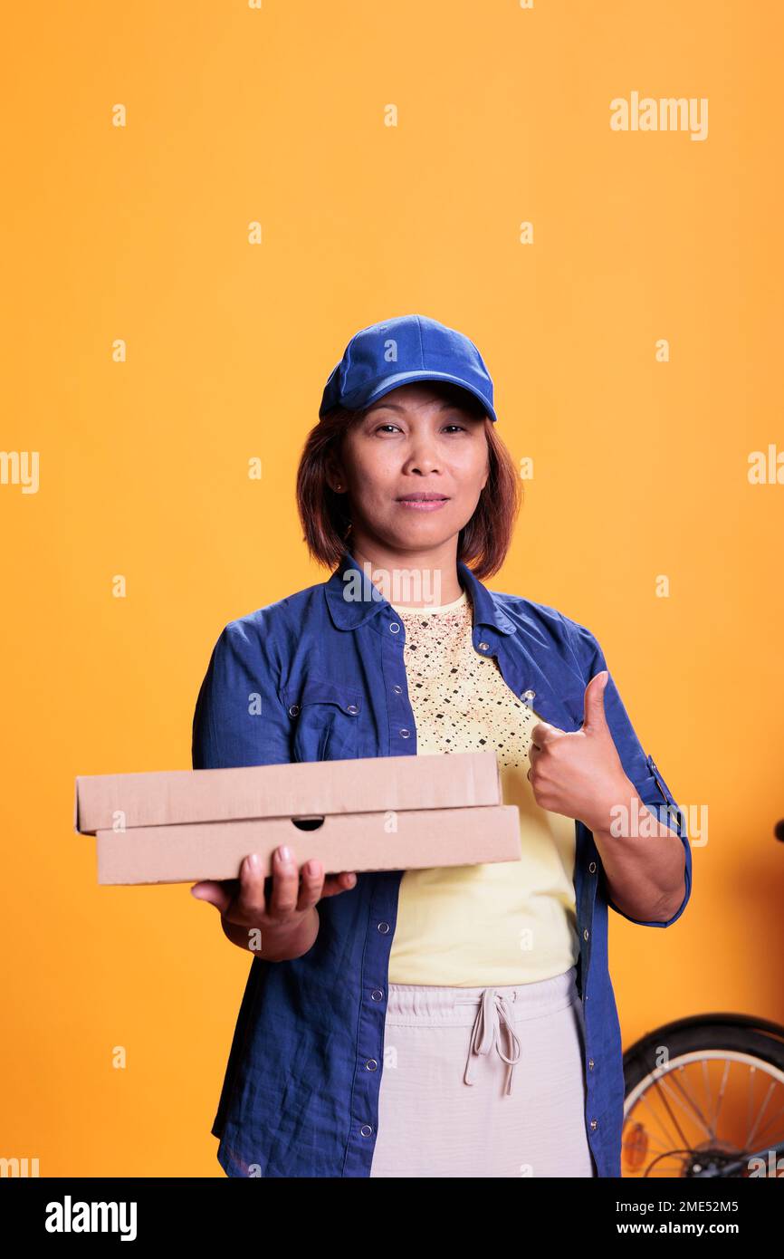 Un employé de la pizzeria asiatique fait des gestes tout en livrant une boîte à plat en carton avec une pizza aux clients. Employé de restaurant apportant des commandes avec vélo. Service de restauration à emporter et concept Banque D'Images