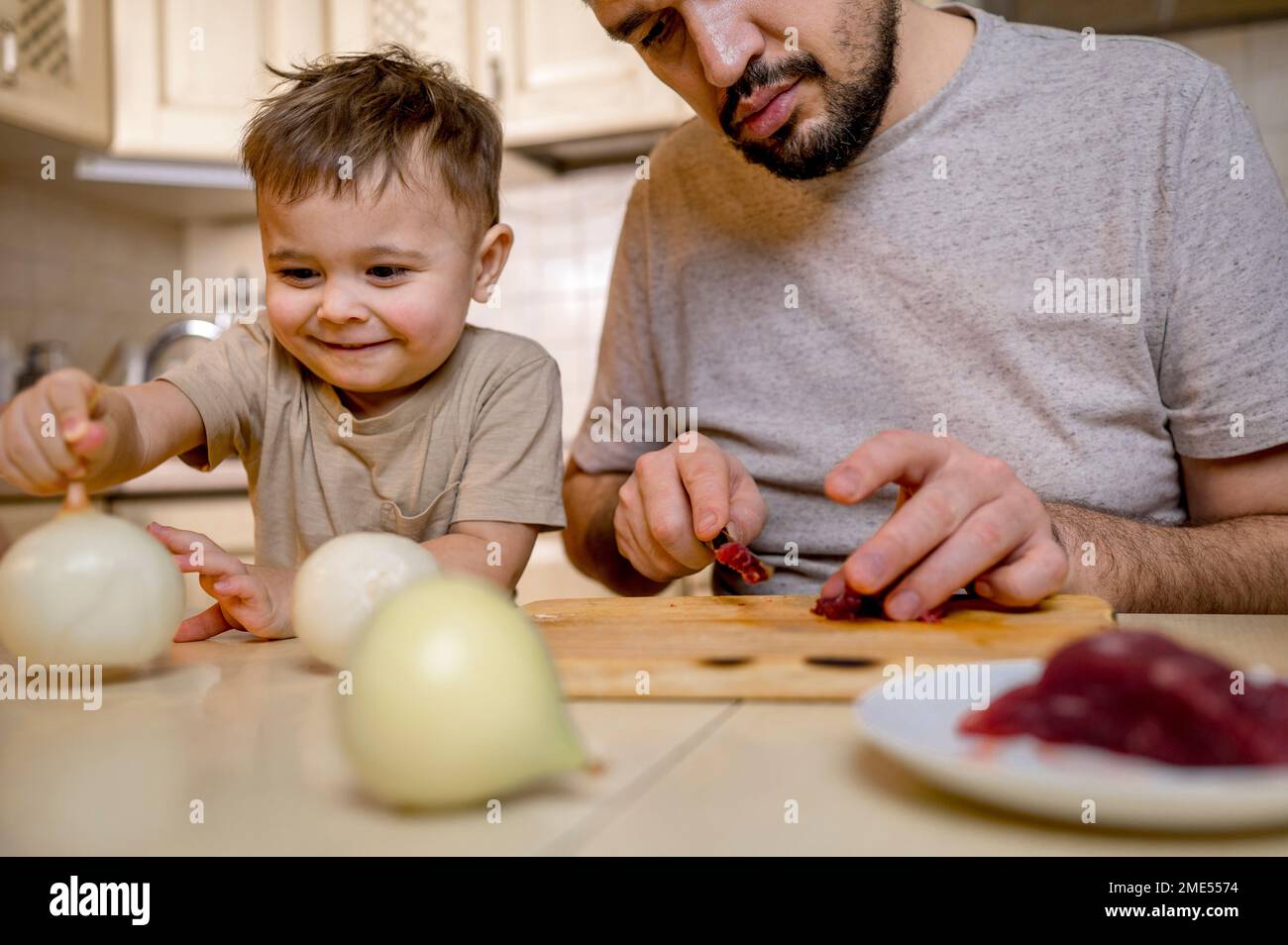 Garçon souriant prenant l'oignon blanc par le père coupant de la viande rouge dans la cuisine Banque D'Images
