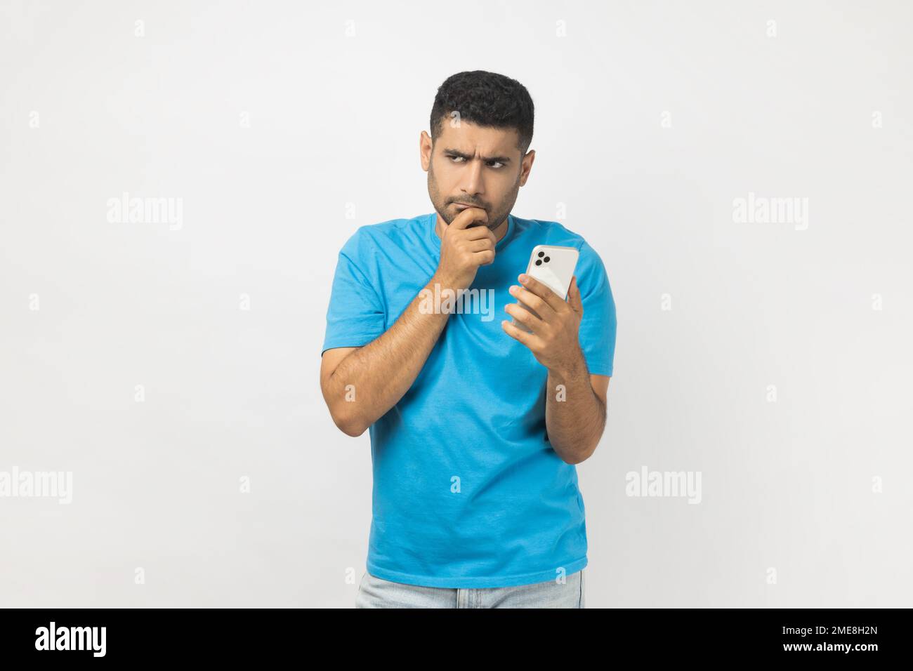 Portrait d'un homme pensif non rasé blogueur portant un T-shirt bleu debout en utilisant le téléphone cellulaire, écrire poste, penser à répondre au message. Prise de vue en studio isolée sur fond gris. Banque D'Images
