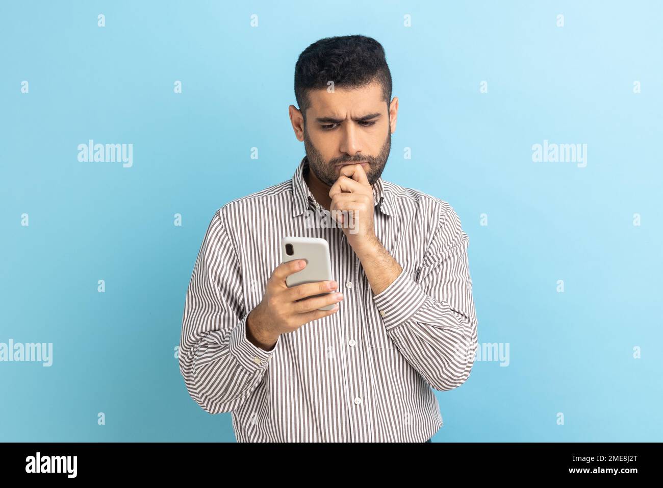 Homme d'affaires sérieux tenant le menton avec un smartphone à la main, envisageant de mettre à jour le logiciel, choisissant des tarifs appropriés, portant une chemise rayée. Studio d'intérieur isolé sur fond bleu. Banque D'Images