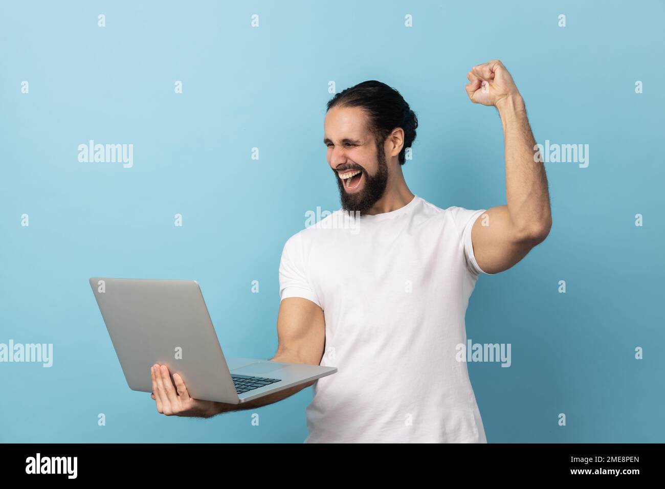 Portrait d'un homme réussi avec une barbe portant un T-shirt blanc criant avec joie et tenant un ordinateur portable, réjouissant la victoire, Paris en ligne. Studio d'intérieur isolé sur fond bleu. Banque D'Images