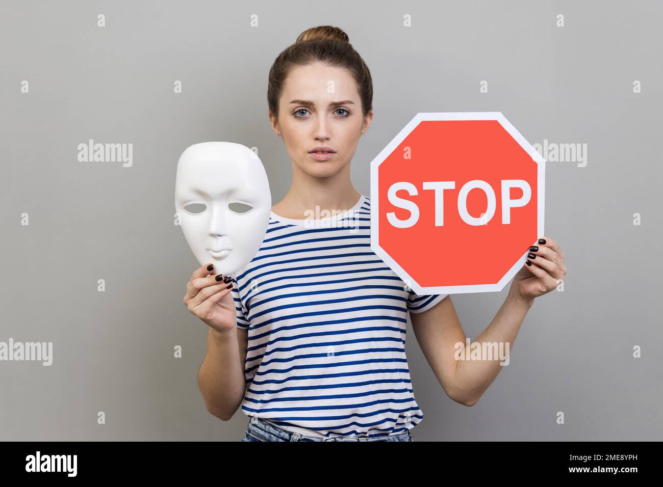 Portrait d'une femme sérieuse portant un T-shirt rayé tenant un masque blanc avec un visage inconnu et un panneau de signalisation rouge, regardant l'appareil photo. Prise de vue en studio isolée sur fond gris. Banque D'Images