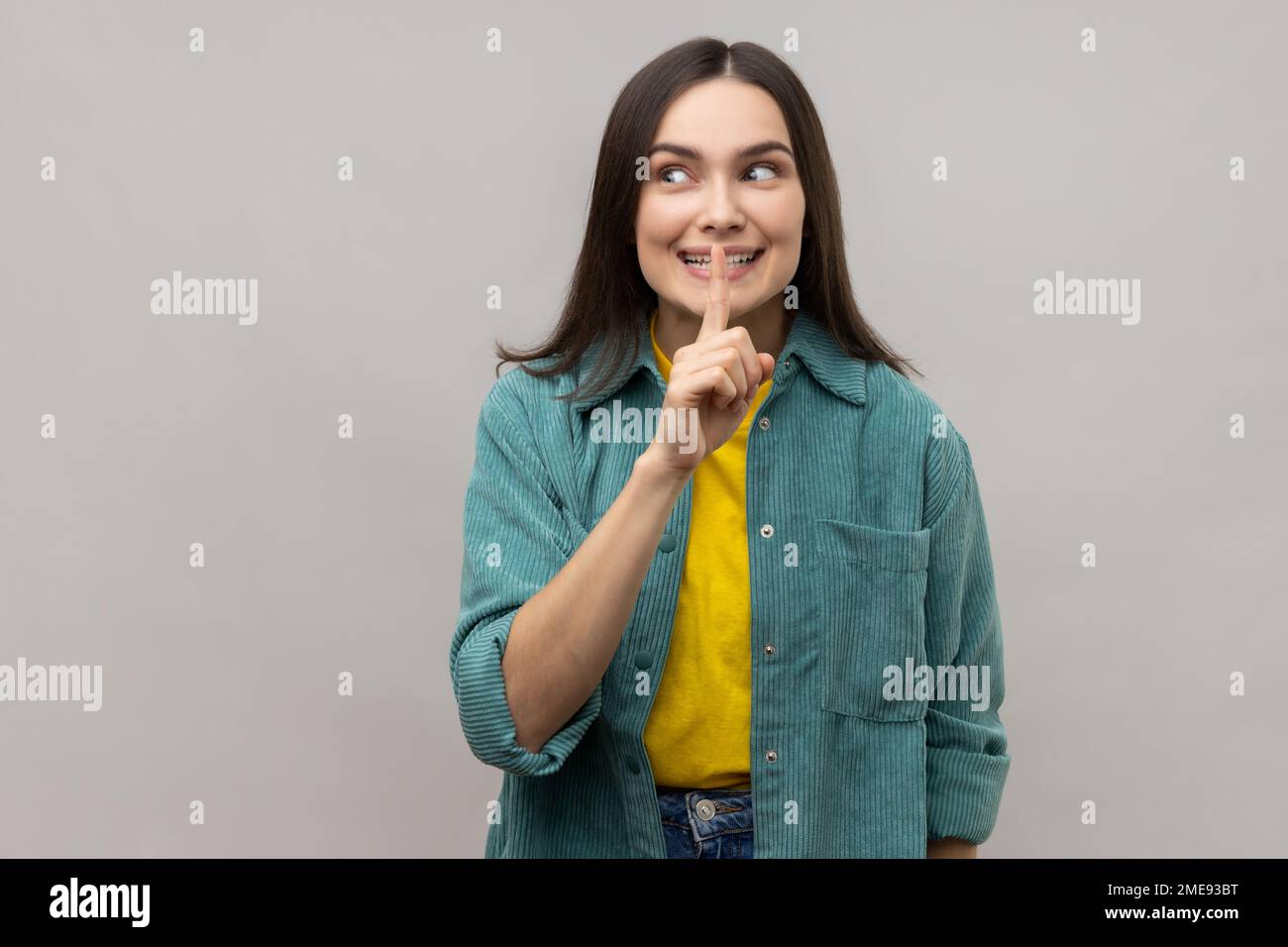 Une femme positive se précipitant avec un geste de silence, tenant le doigt sur les lèvres et souriant à l'appareil photo, demandant de garder le secret, portant une veste de style décontracté. Prise de vue en studio isolée sur fond gris. Banque D'Images