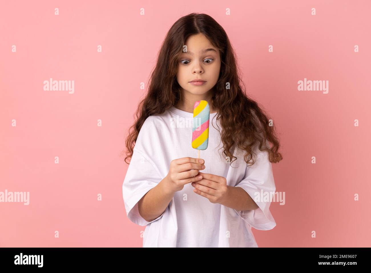 Portrait de la petite fille surprise choquée portant un T-shirt blanc tenant une délicieuse crème glacée, regarde le dessert avec une expression étonnante. Studio d'intérieur isolé sur fond rose. Banque D'Images