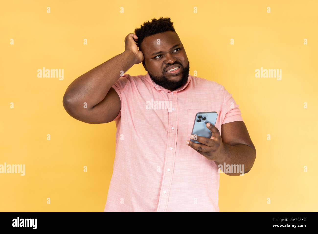 Portrait d'un homme pensif à barbe portant une chemise rose debout avec un téléphone portable, touchant la tête, doutant sentiment de suspicion. Studio d'intérieur isolé sur fond jaune. Banque D'Images