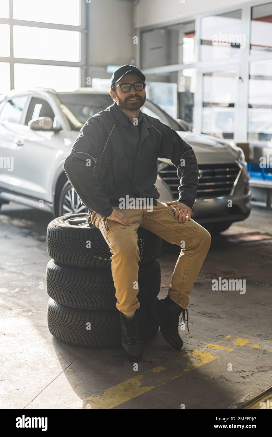 Photo verticale. Prise de vue complète. Mécanicien barbu avec des lunettes assis sur une pile de pneus et souriant à la caméra. Voiture en arrière-plan. Concept atelier de réparation. Photo de haute qualité Banque D'Images