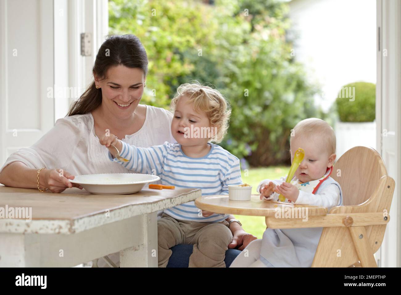 Femme assise à une table avec un tout-petit sur les genoux et un bébé en chaise haute, 2 ans 4 mois et 9 mois Banque D'Images
