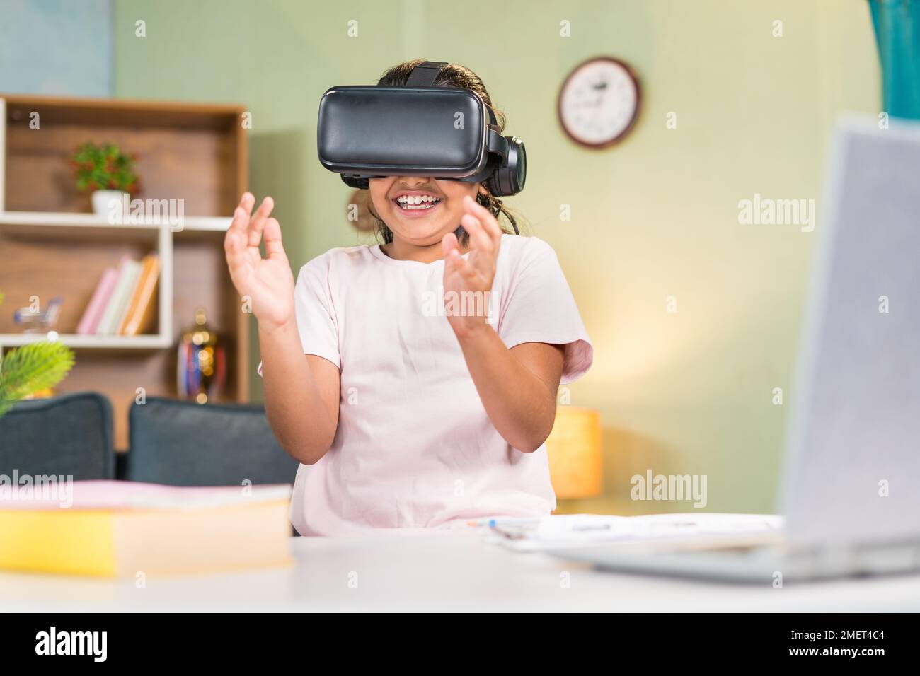 Joyeux adolescent souriant enfant de fille avec vr ou jeu de tête virtuel expérience en ligne ou classe virtuelle en utilisant le geste de la main devant la table d'étude à la maison - Banque D'Images