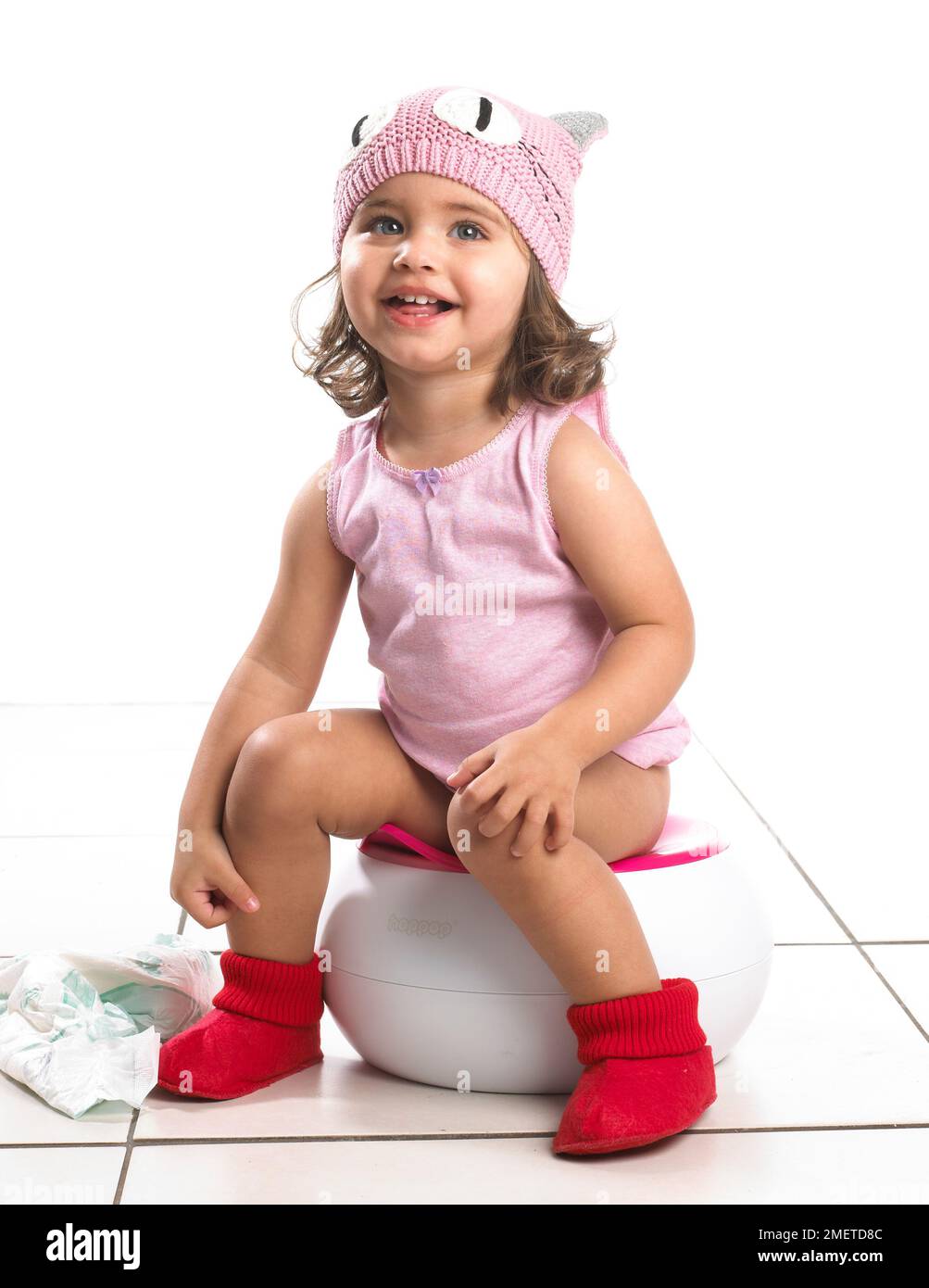 Fille portant un gilet rose et un chapeau en laine rose, des pantoufles rouges assis sur un pot blanc, couche sur le sol, 20 mois Banque D'Images