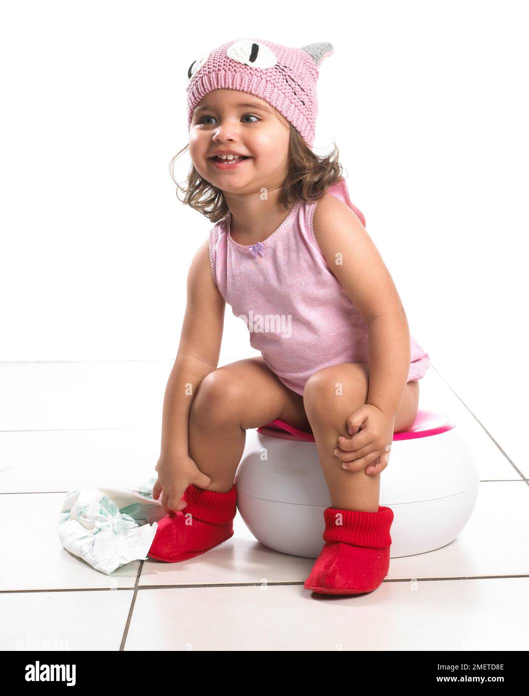Fille portant un gilet rose et un chapeau de laine rose, des pantoufles rouges assis sur un pot blanc tenant la neppy, 20 mois Banque D'Images
