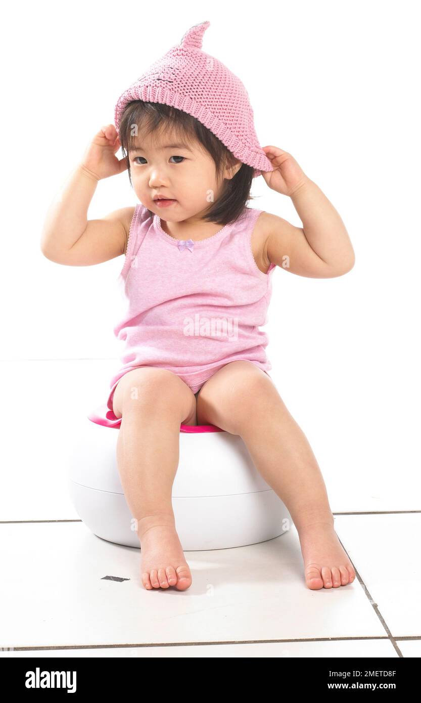 Fille portant un gilet rose assis sur un pot blanc, portant un chapeau de laine rose, 20 mois Banque D'Images
