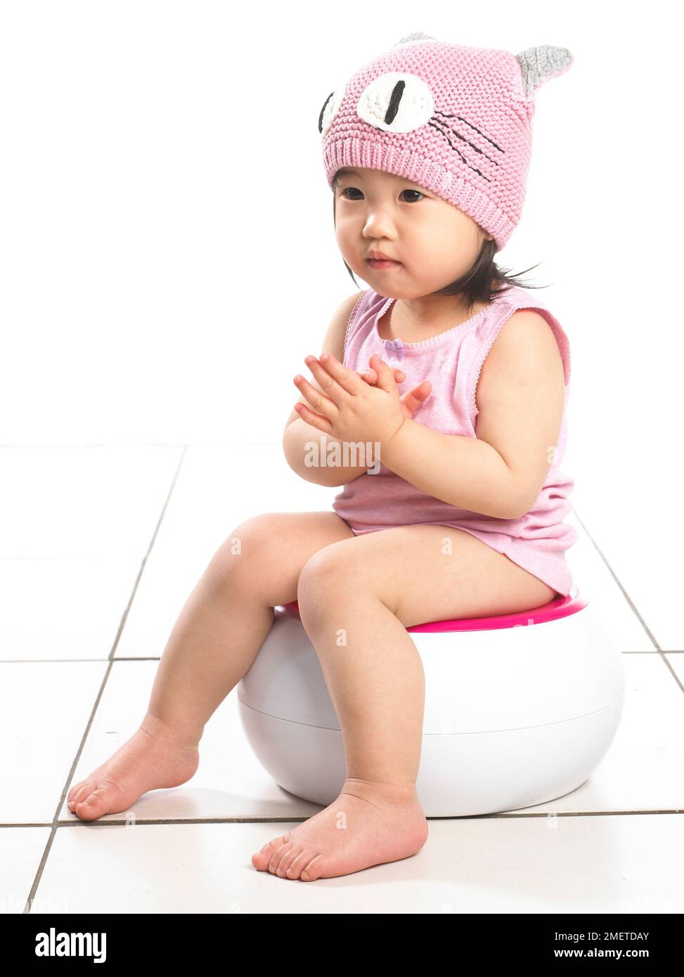 Fille portant un gilet rose et un chapeau en laine rose, assise sur un pot blanc, 20 mois Banque D'Images