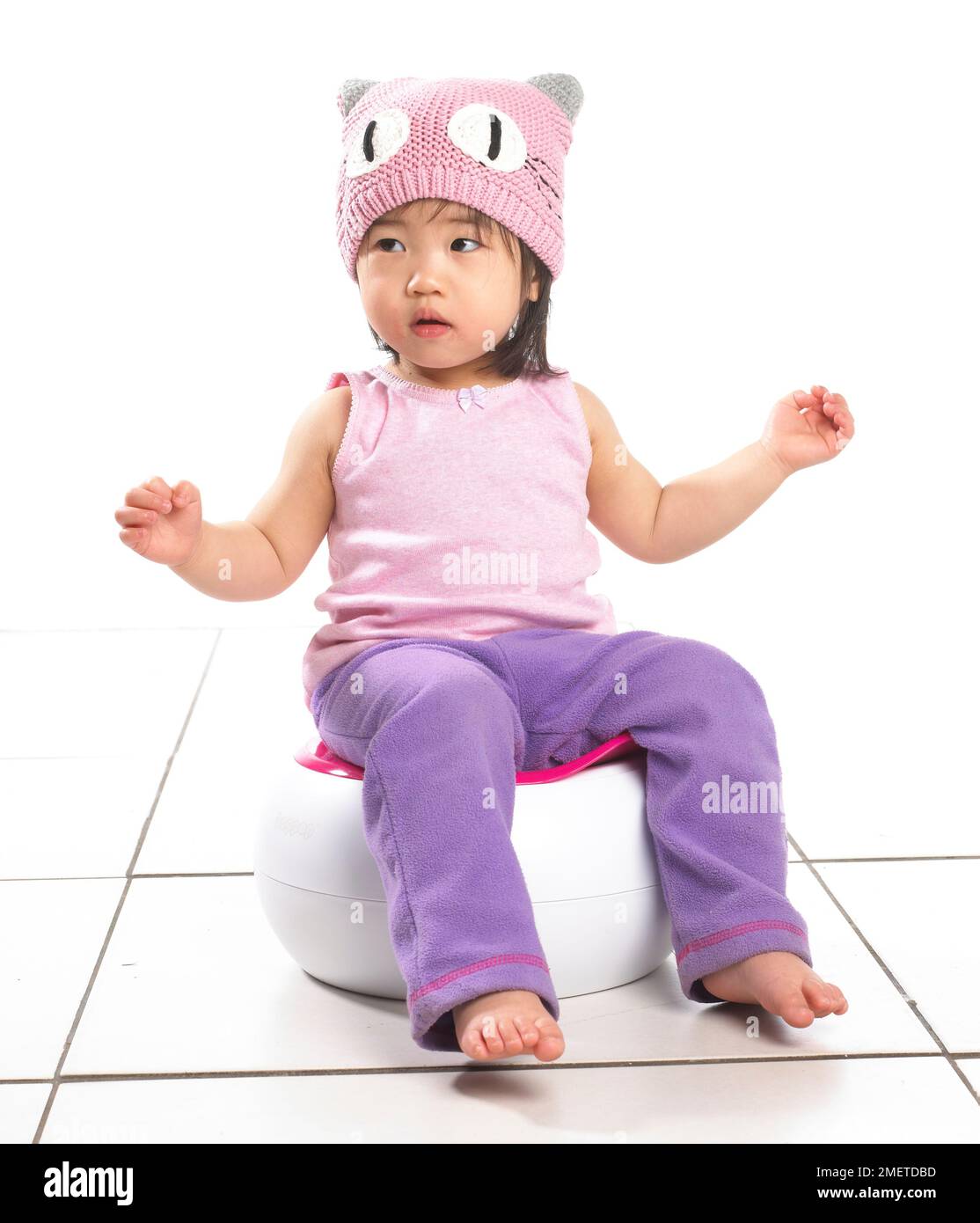 Fille portant un gilet rose, un chapeau de laine rose et un pantalon de jogging violet assis sur un pot blanc, 20 mois Banque D'Images