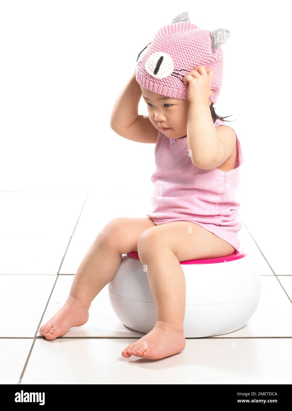Fille portant le gilet rose assis sur pot blanc, mettant sur chapeau de laine rose, 20 mois Banque D'Images