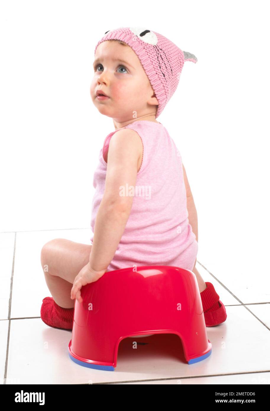 Fille assise sur pot rouge regardant derrière elle, portant chapeau de laine rose, vue arrière, 19 mois Banque D'Images