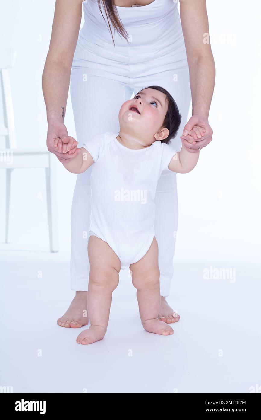 Bébé fille (40 semaines) debout, regardant la mère qui se tient derrière elle et tient ses mains Banque D'Images