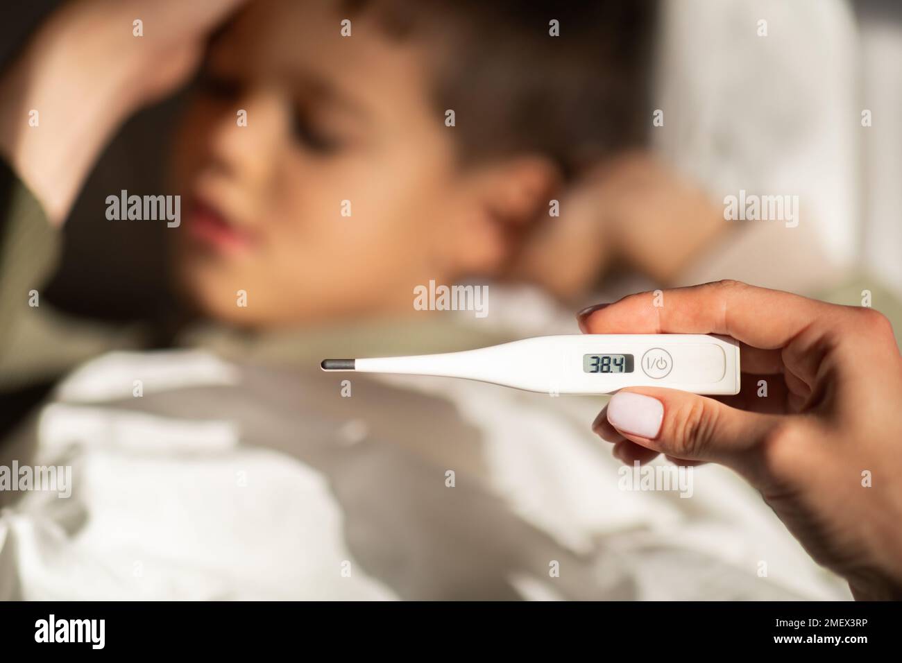 Un petit garçon européen fatigué se trouve sur le lit, dormant, maman mesure la température avec un thermomètre dans la chambre Banque D'Images