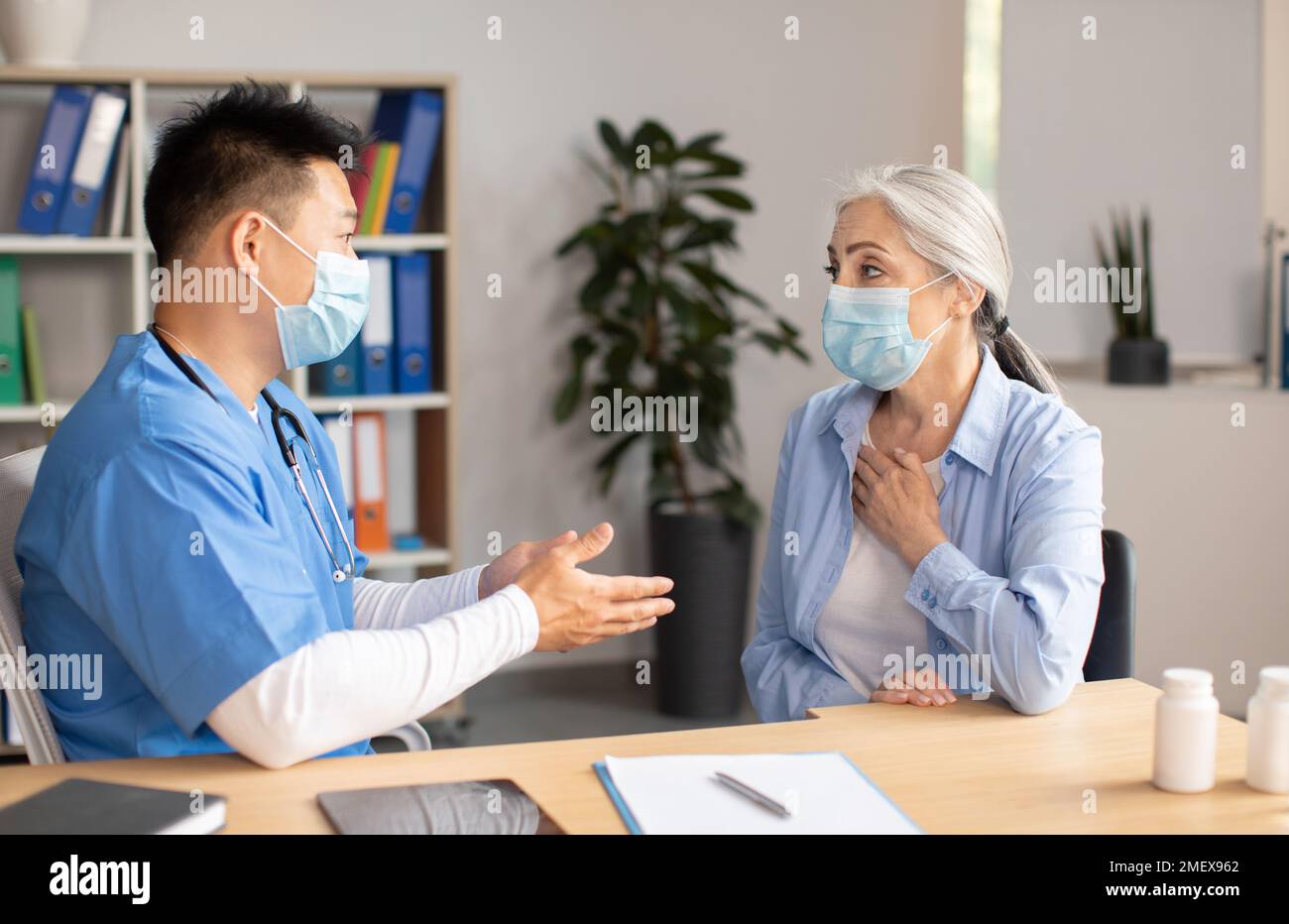 Un médecin coréen sérieux et mature dans un masque de protection consulte une patiente européenne âgée en clinique Banque D'Images