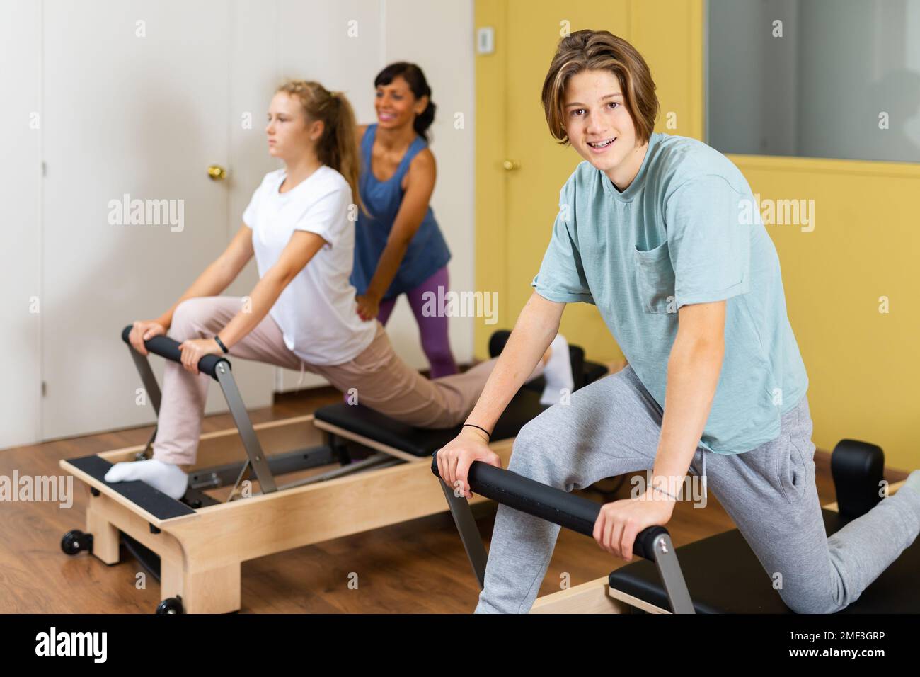 Jeune garçon regardant dans l'appareil photo tout en s'entraîner pilates dans la salle de gym. Leur entraîneur hispanique femme aidant la fille avec des exercices sur les pilates reformer en arrière-plan. Banque D'Images