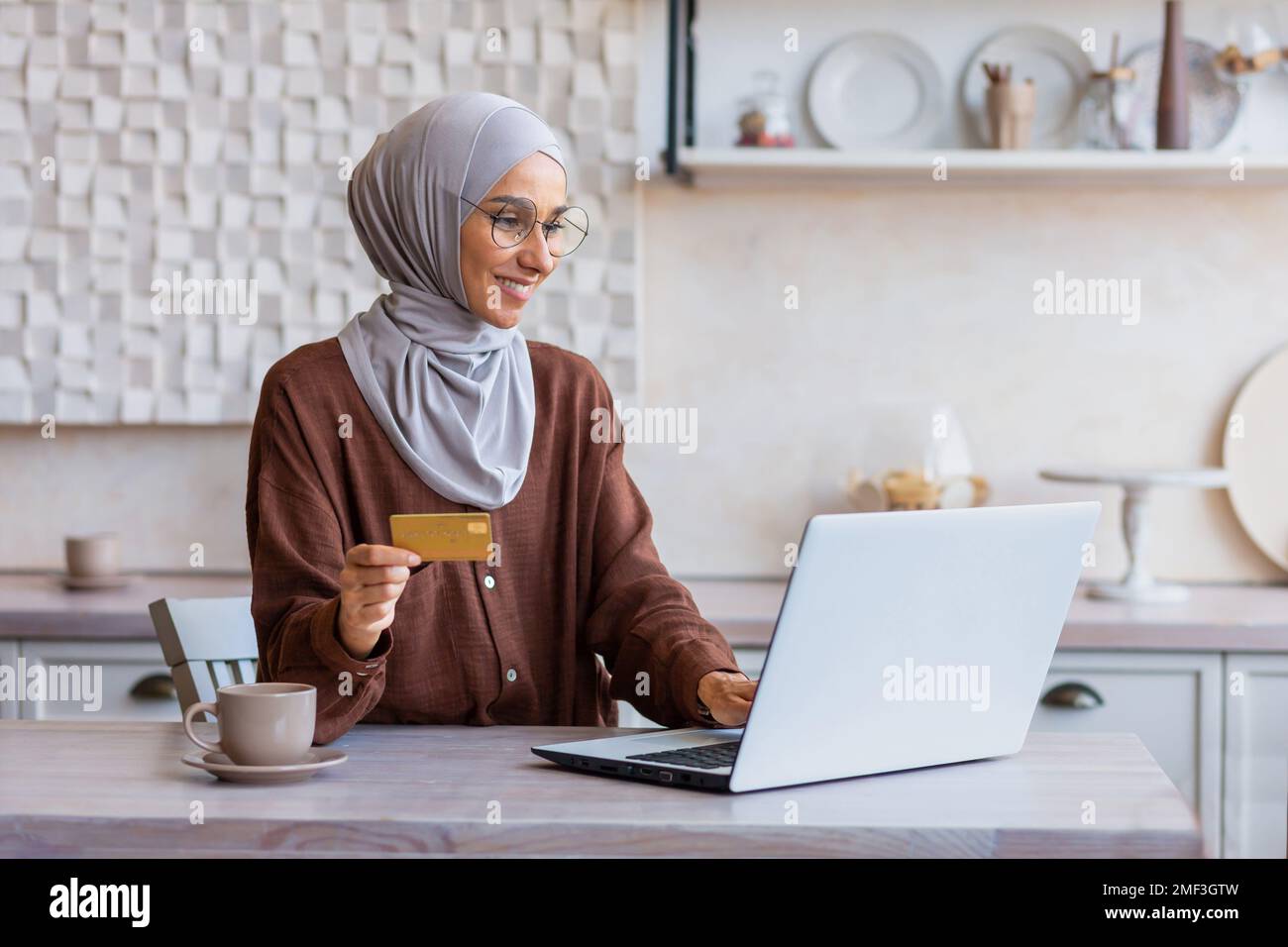 Femme gaie et réussie dans hijab à la maison dans la cuisine avec  ordinateur portable et carte de crédit bancaire fait l'achat en ligne dans  la boutique en ligne, la femme musulmane