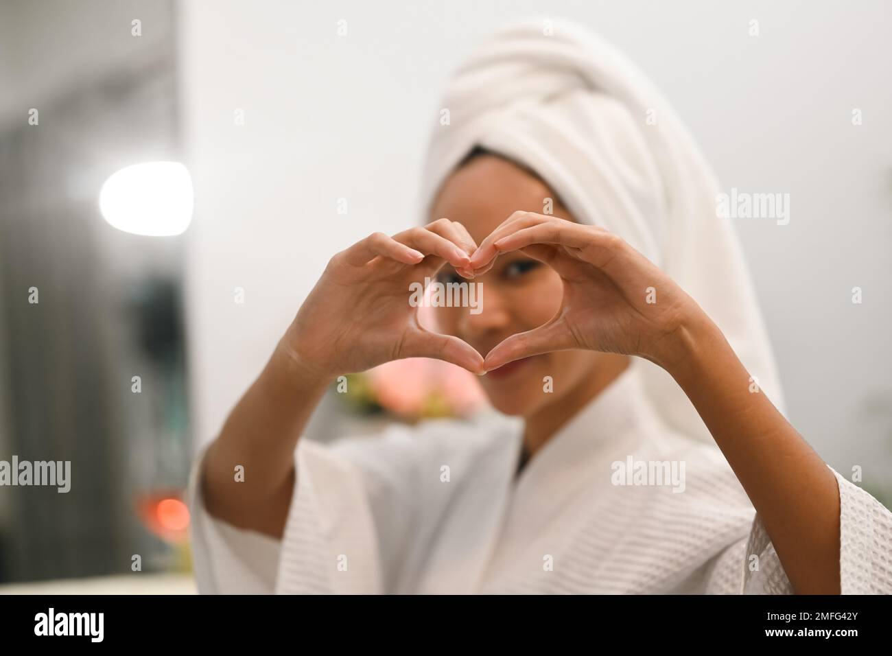 Jeune femme asiatique faisant le symbole de coeur forme avec les mains souriant à son reflet dans le miroir. Concept de beauté et d'auto-soins Banque D'Images